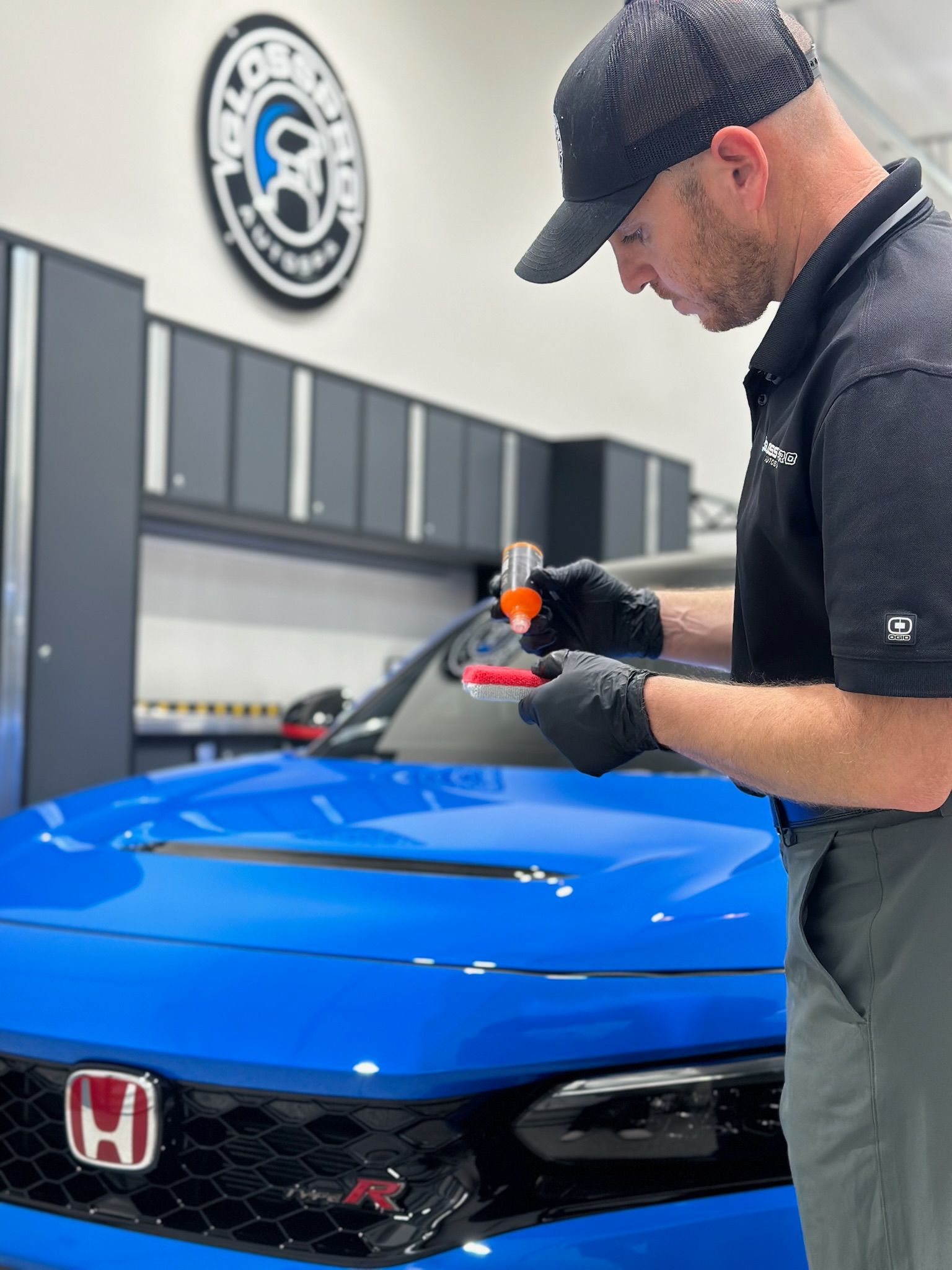 A man in a cap and gloves applies a liquid to a red applicator on the hood of a blue car, inside a well-lit shop.