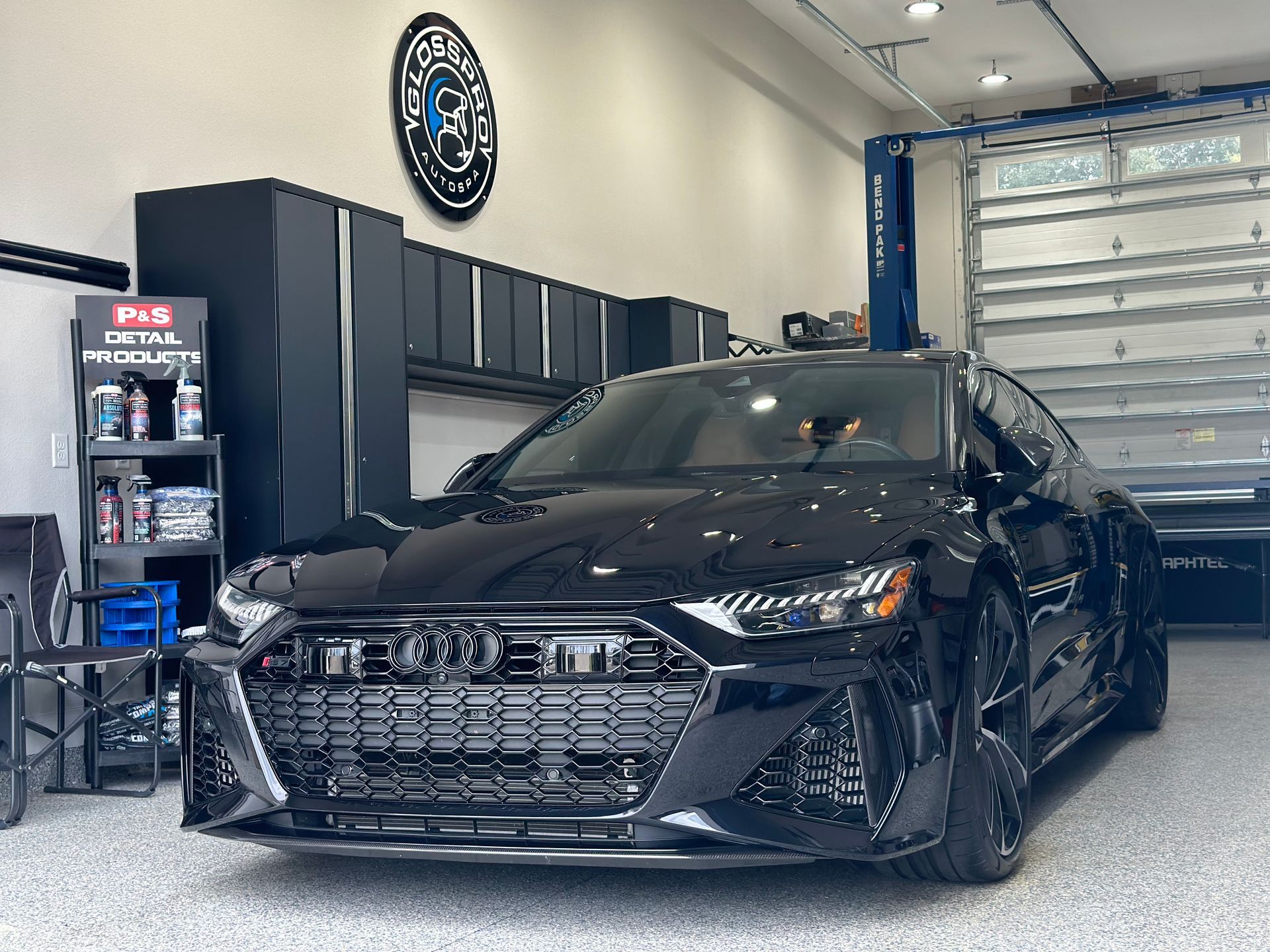 A shiny black Audi RS7 parked inside a garage. The car faces the camera; the garage has black cabinets and a lift.