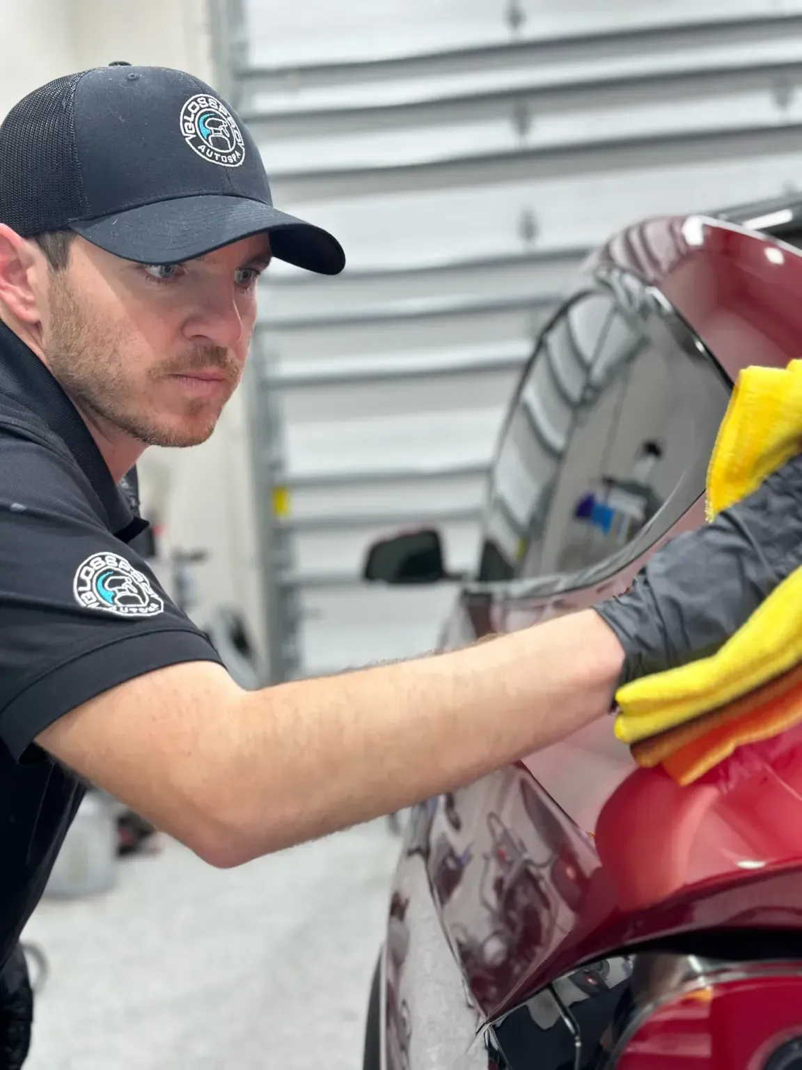 A man wearing a cap and gloves wipes a red car with a yellow cloth in a garage. He looks focused.