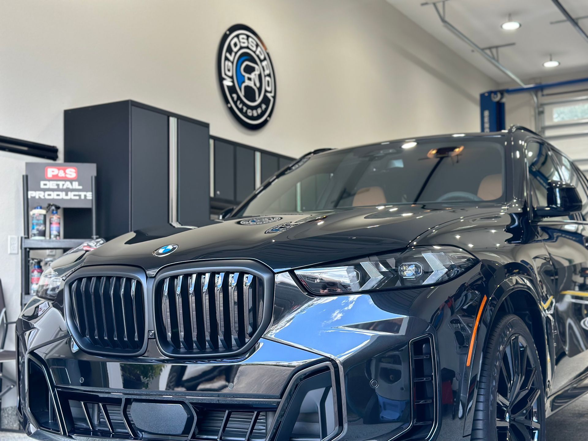Black BMW SUV in a garage, reflecting surrounding objects, with a company logo on the wall and detailing products visible.