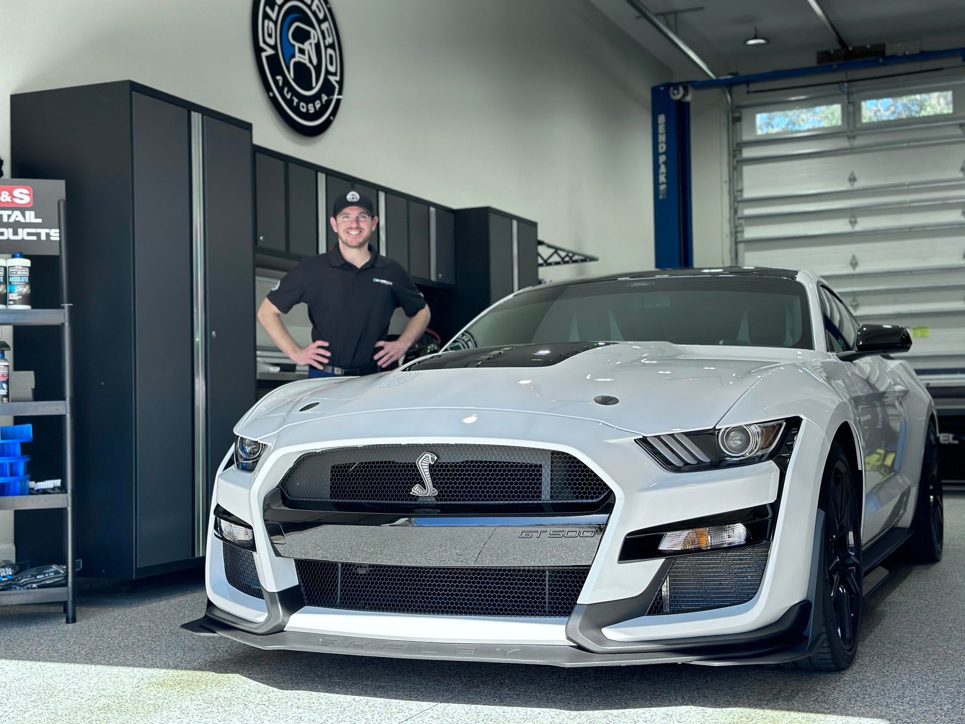 Man in a black shirt stands next to a white Ford Mustang in a garage.