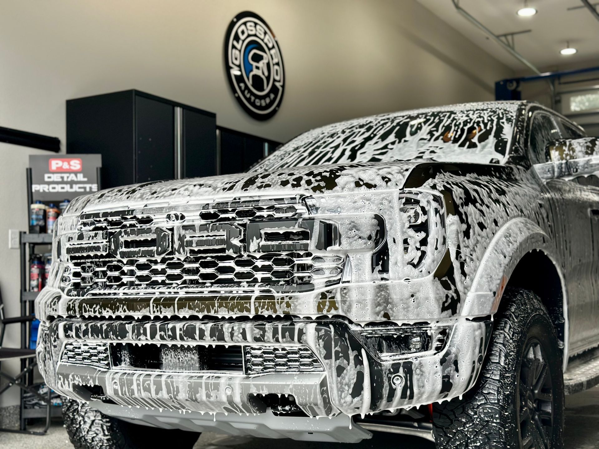 A black truck covered in white foam, likely for washing, with a detailing shop interior and logo in the background.