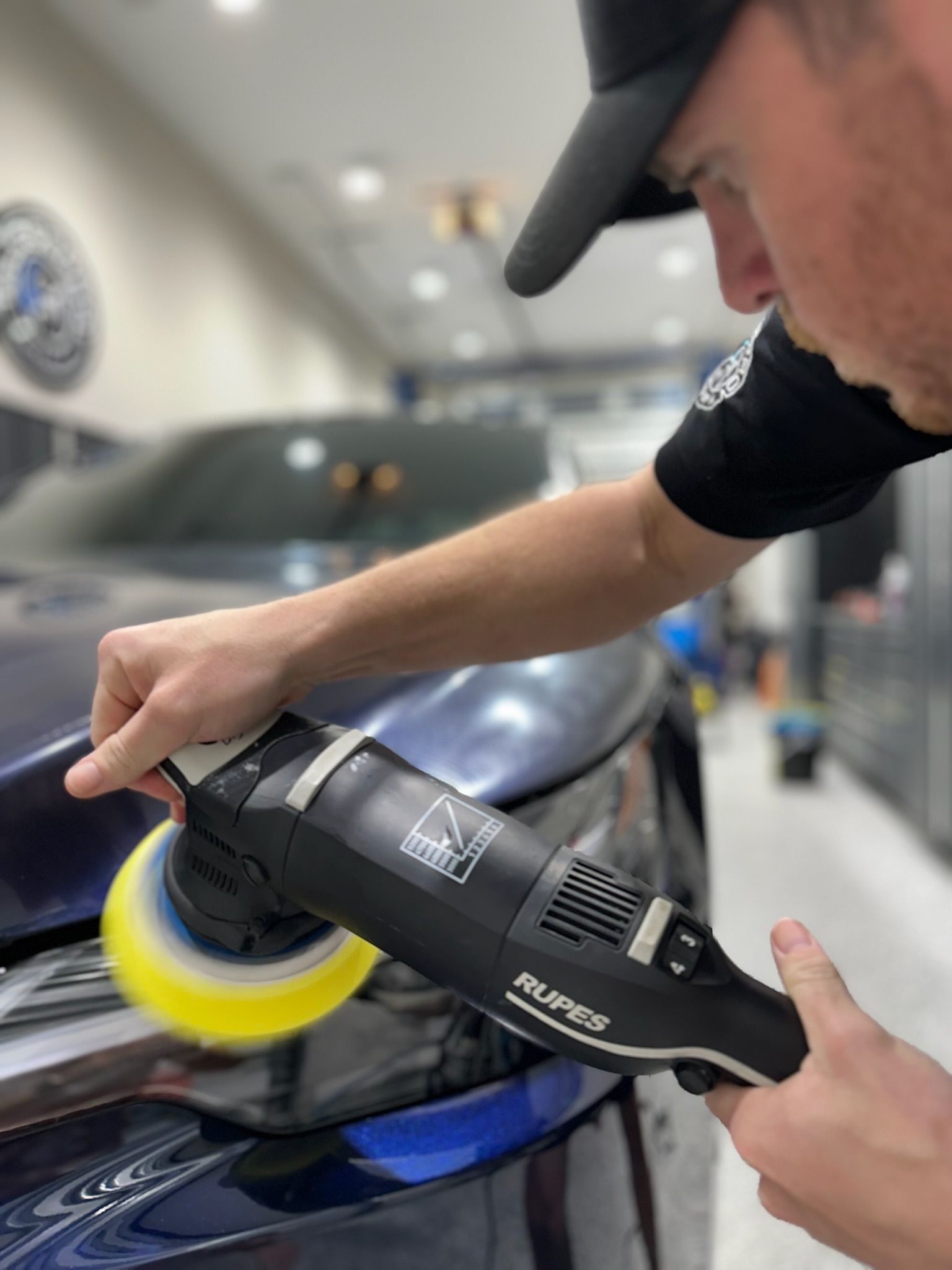 A person polishes a blue car hood with a Rupes polisher in a detail shop. The person wears a black cap and shirt.