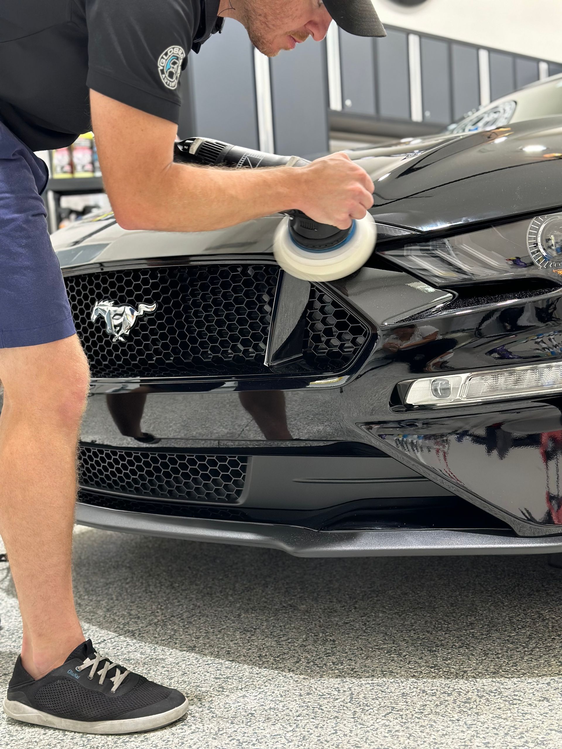 A person polishing a black Ford Mustang. They are wearing a black shirt, blue shorts, and a cap, working in a garage.