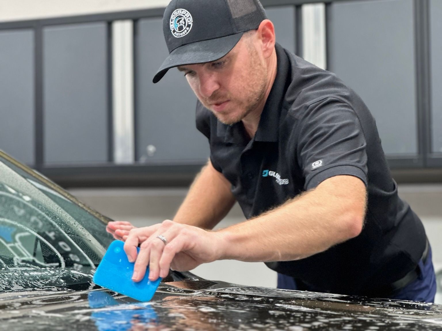 A man in a black cap and shirt applies a blue squeegee to the hood of a car, likely applying tint or protective film.