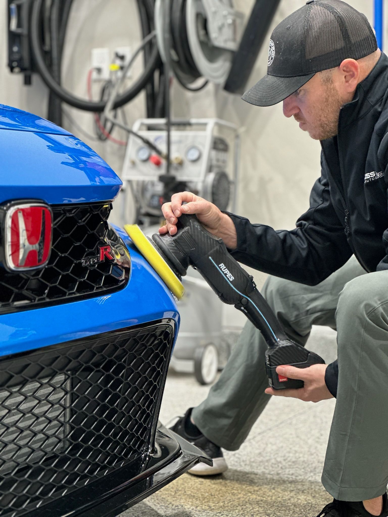 A man polishes a blue Honda car bumper with a buffer in a garage setting. He wears a black cap and jacket.