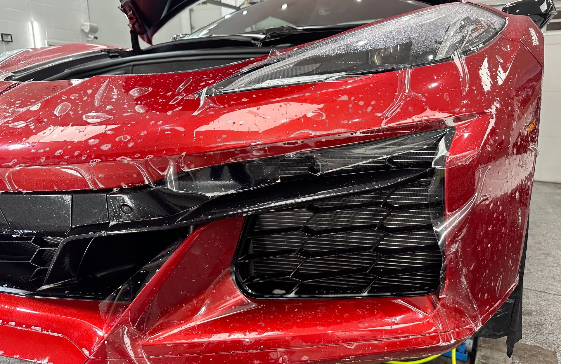 Red sports car front bumper with protective film being applied. Detail of black grill and headlight area.