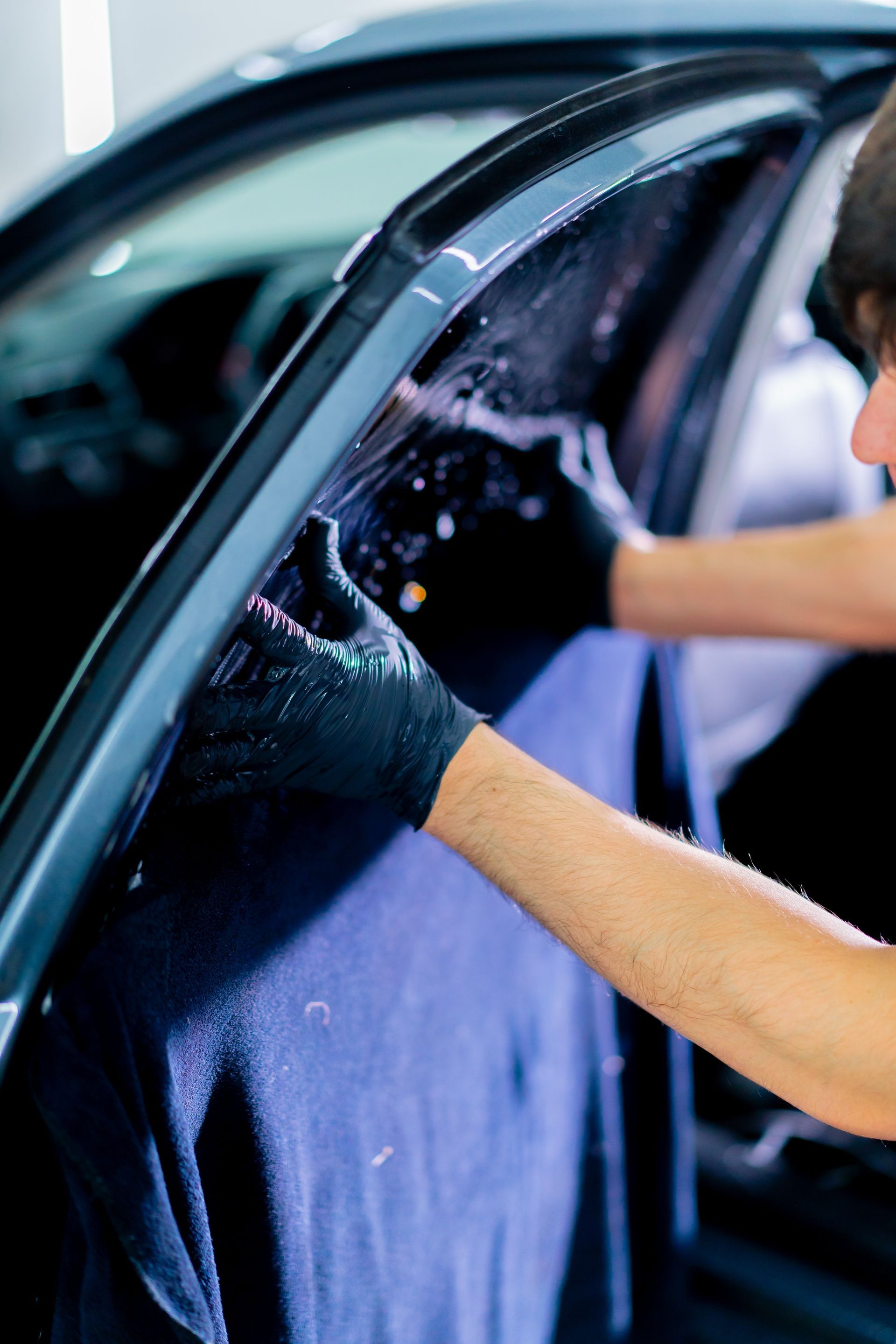 A person in black gloves applying tinted film to a car window, using a spray bottle and blue towel.