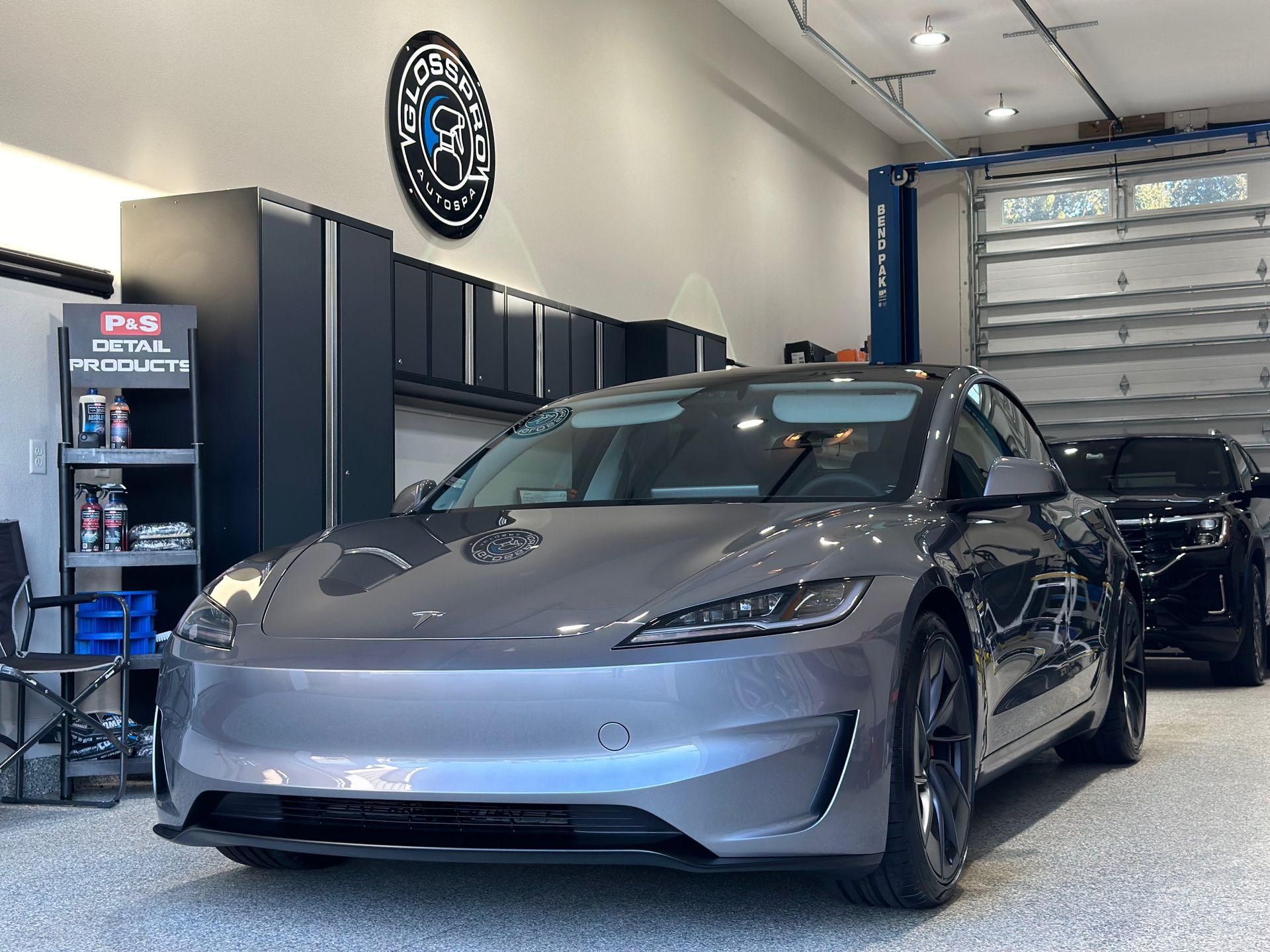 A gray Tesla sedan in a garage, another black car is in the background. The garage has cabinets and car detailing products.