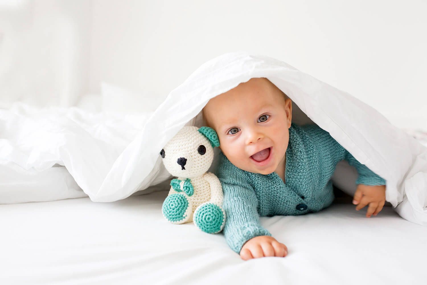 Smiling baby in a blue sweater, peeking from under white blanket with a teddy bear.