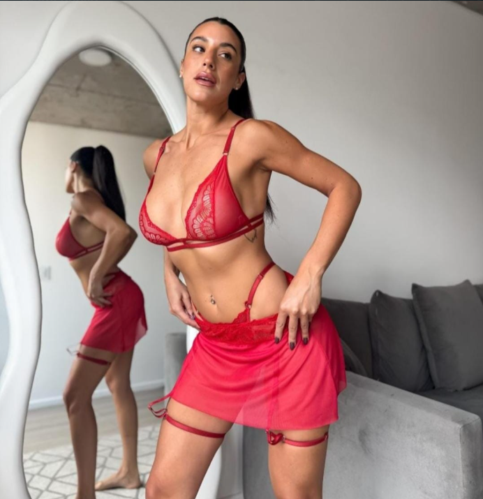 Mujer en lencería roja posando. Está en un interior con un espejo y un sofá visibles.