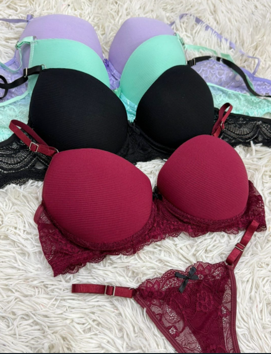 Sujetadores de varios colores (violeta, verde, negro, burdeos) apilados sobre una superficie peluda blanca; un conjunto incluye ropa interior a juego.