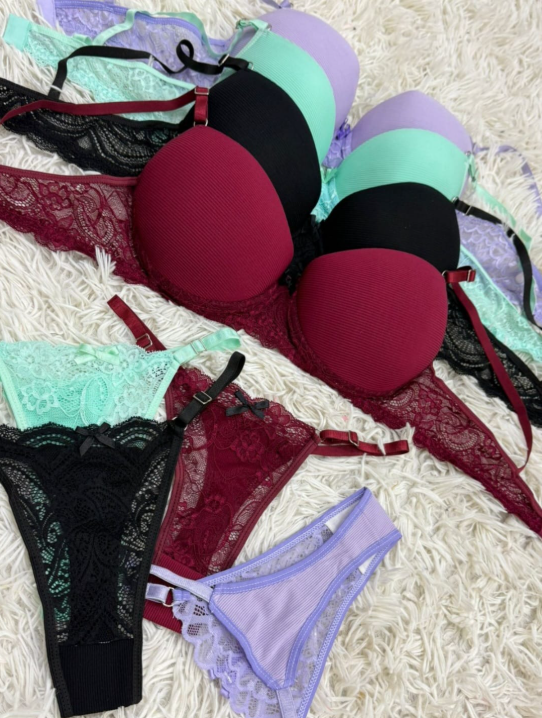 Sujetadores de varios colores (violeta, verde, negro, burdeos) apilados sobre una superficie peluda blanca; un conjunto incluye ropa interior a juego.