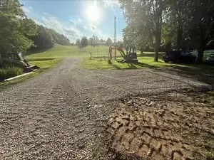 Gravel driveway leading to a sunny park with trees and a playground in the distance