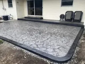 Stamped concrete patio with black edging beside a house and sliding glass door, with outdoor chairs on the right