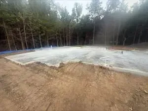Gravel foundation pad beside a wooded tree line under an overcast sky