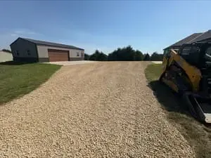 Gravel driveway leading to a garage, with a skid steer on the right and a grassy yard under a clear sky.