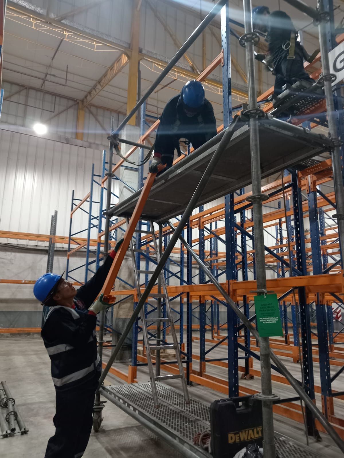 Dos trabajadores construyen andamios dentro de un almacén, uno de ellos intenta alcanzarlos con un palo.