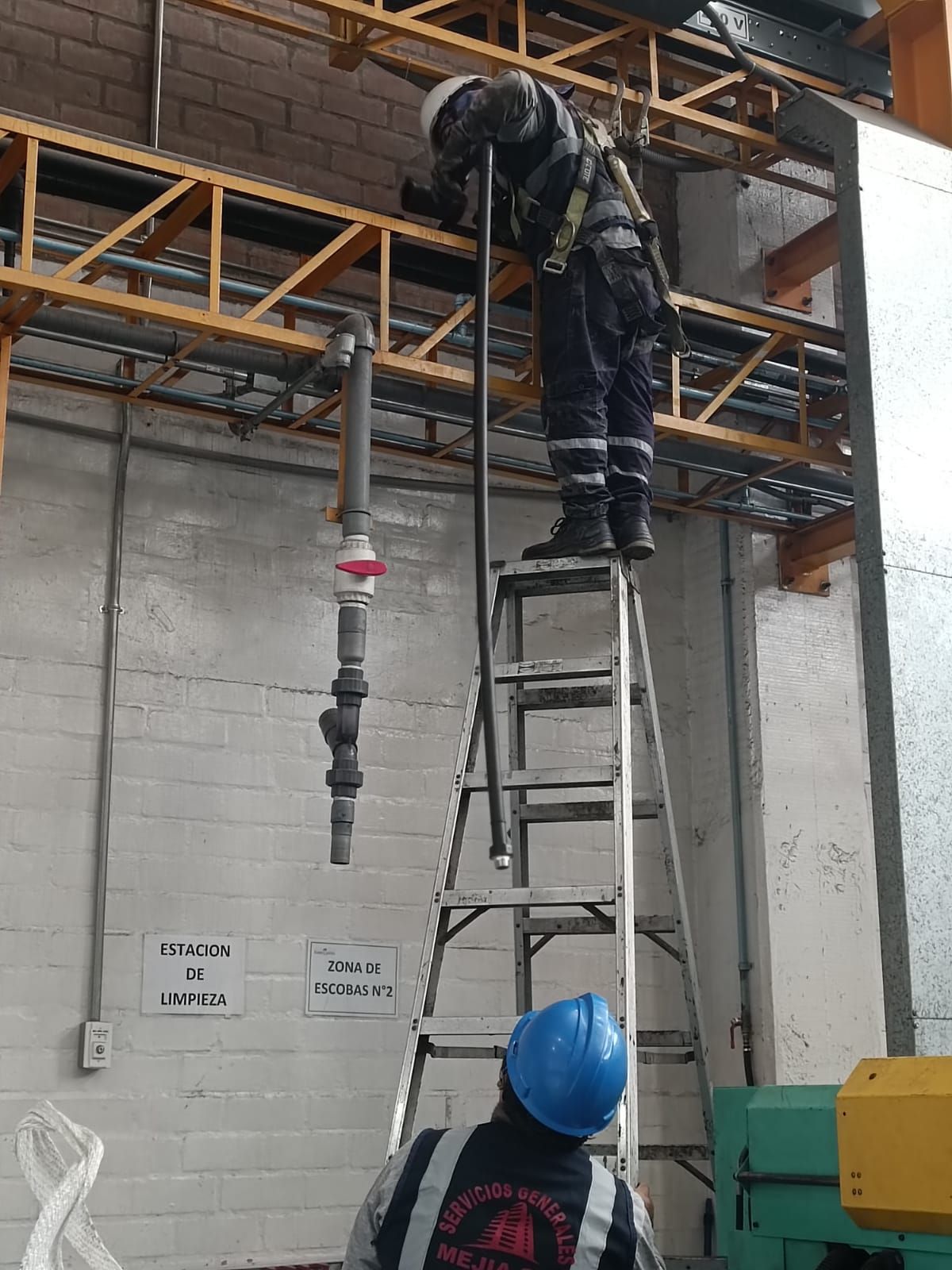 Dos trabajadores en una fábrica; uno en una escalera trabajando en una tubería, otro debajo.