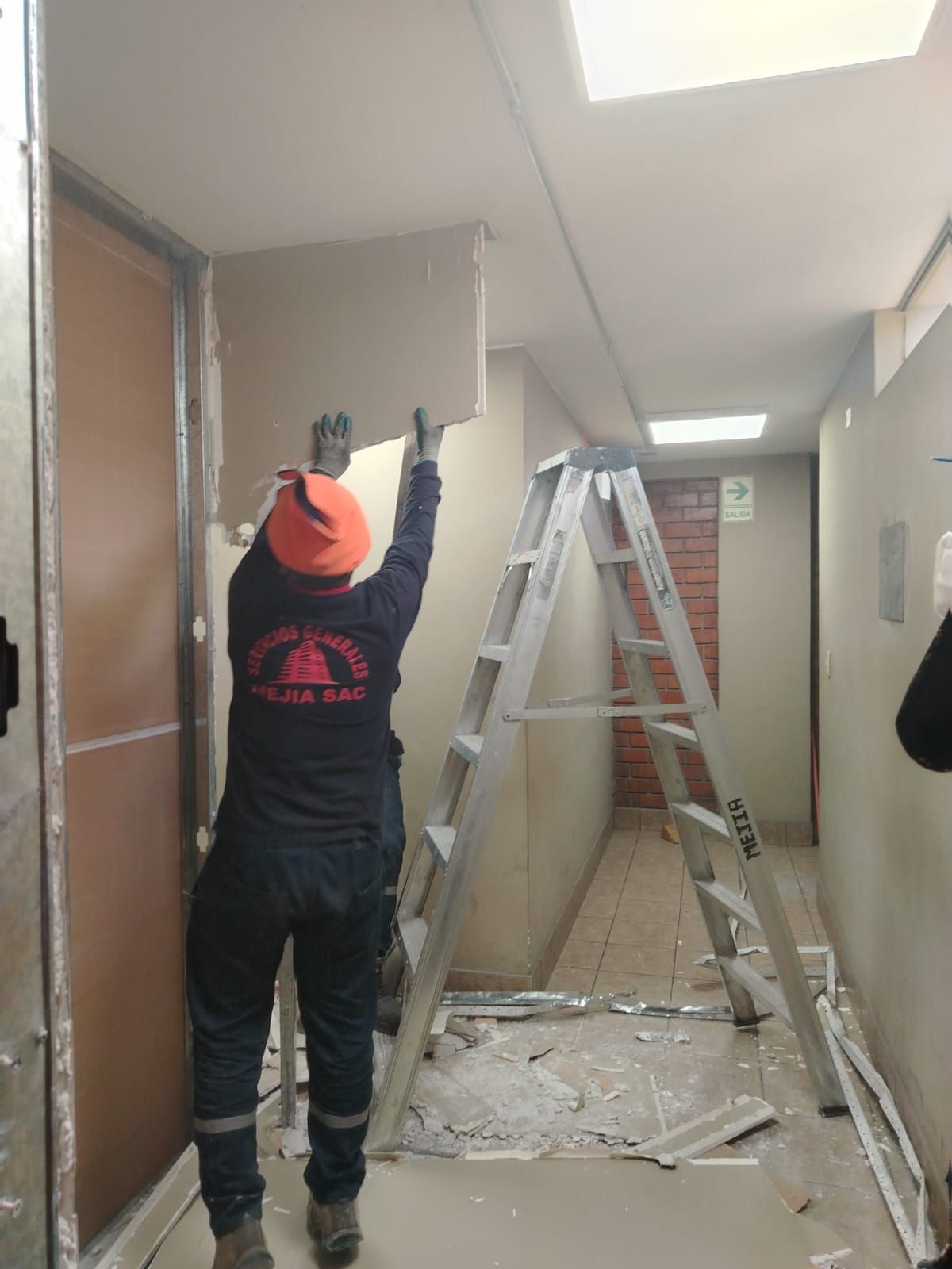 Una persona instala paneles de yeso en un pasillo estrecho. Está subido a una escalera, con un gorro de lana