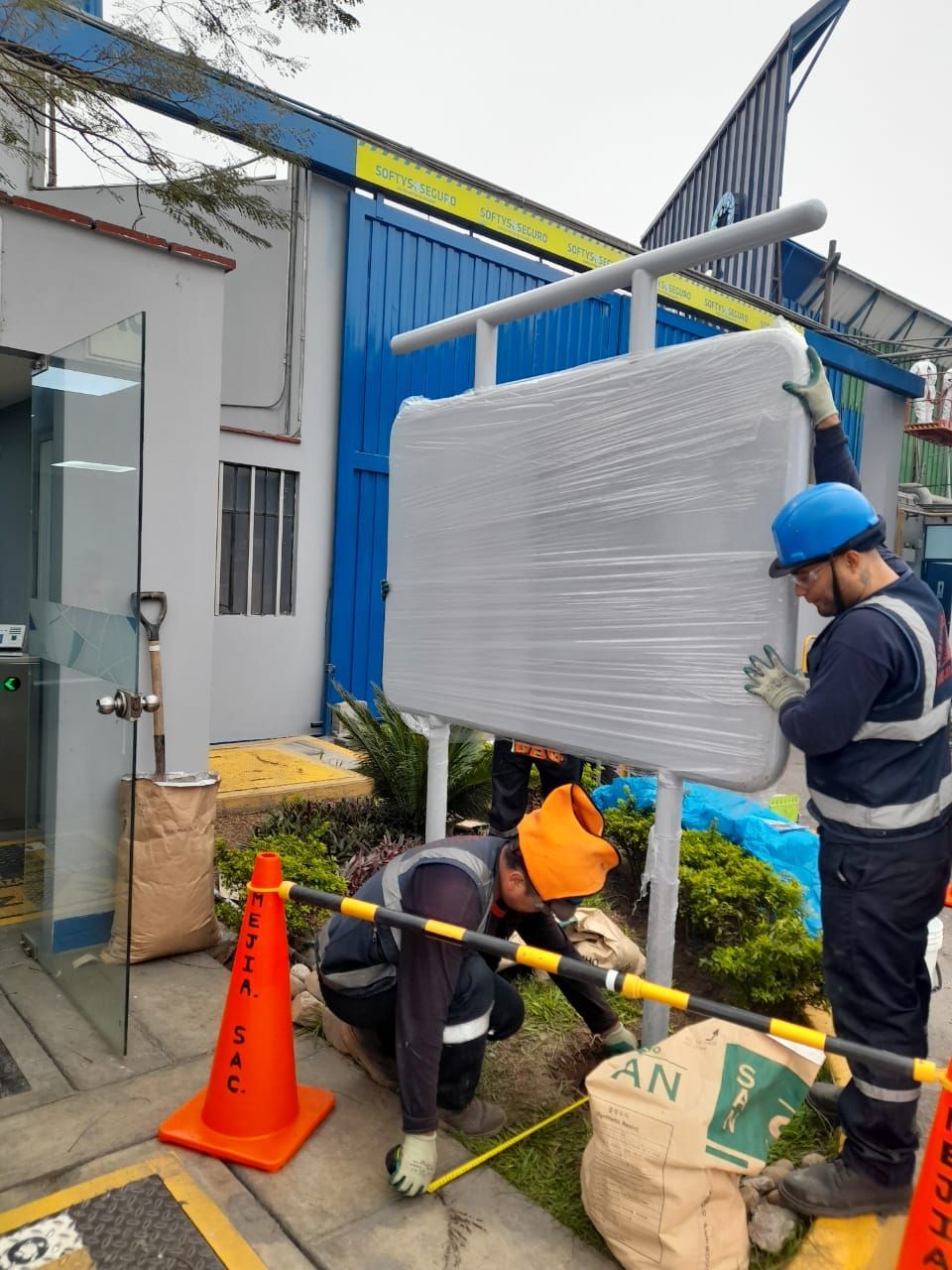 Trabajadores instalando un letrero envuelto en plástico frente a un edificio azul. Uno mide, otro sostiene el letrero.