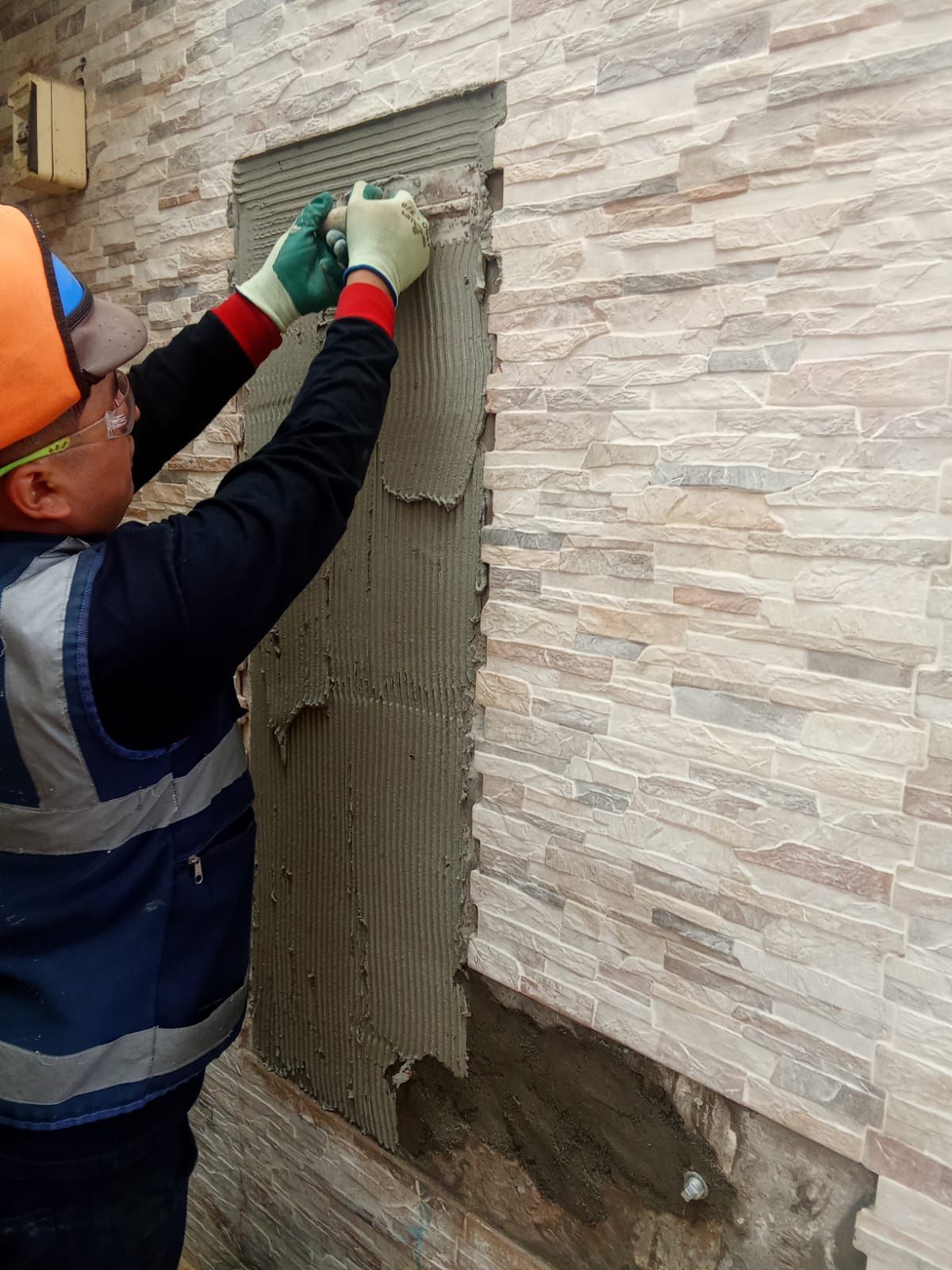 Obrero de la construcción instalando revestimiento de piedra en una pared, usando equipo de seguridad y aplicando mortero.
