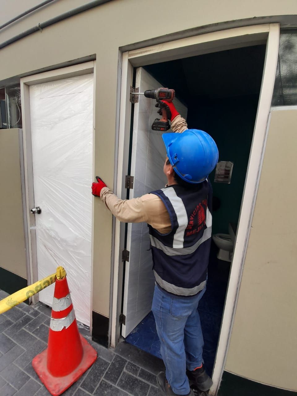 Un trabajador con casco azul instala una puerta con un taladro eléctrico en el exterior. Hay un cono de tráfico cerca.