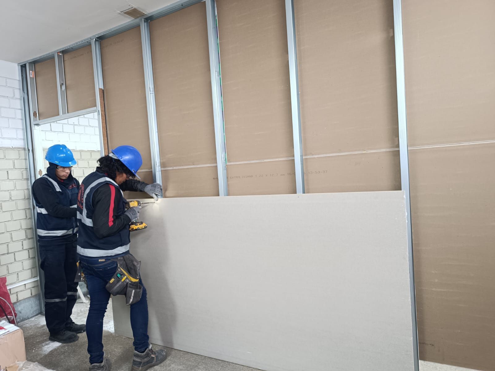 Dos trabajadores instalando paneles de yeso sobre montantes metálicos dentro de una habitación, usando cascos de seguridad.