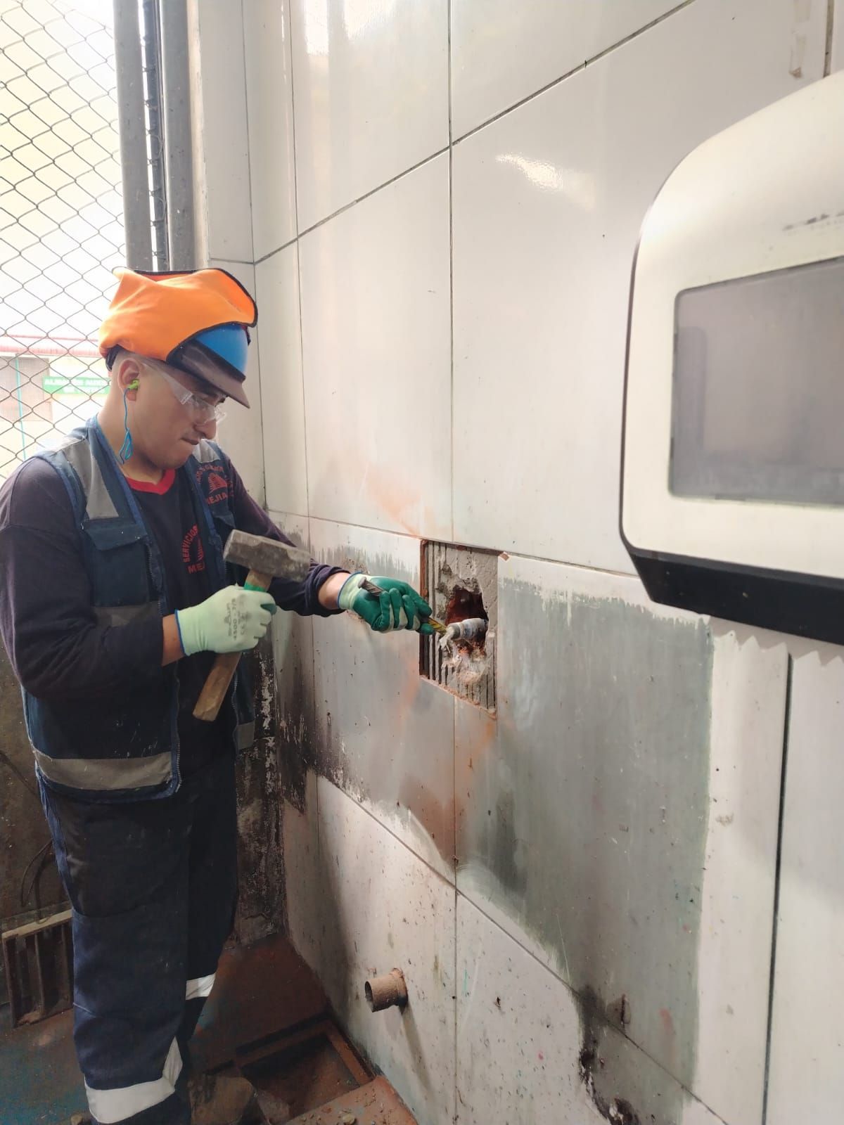 Un trabajador con chaleco de seguridad y casco usa un martillo para romper una pared, al lado de un electrodoméstico blanco.