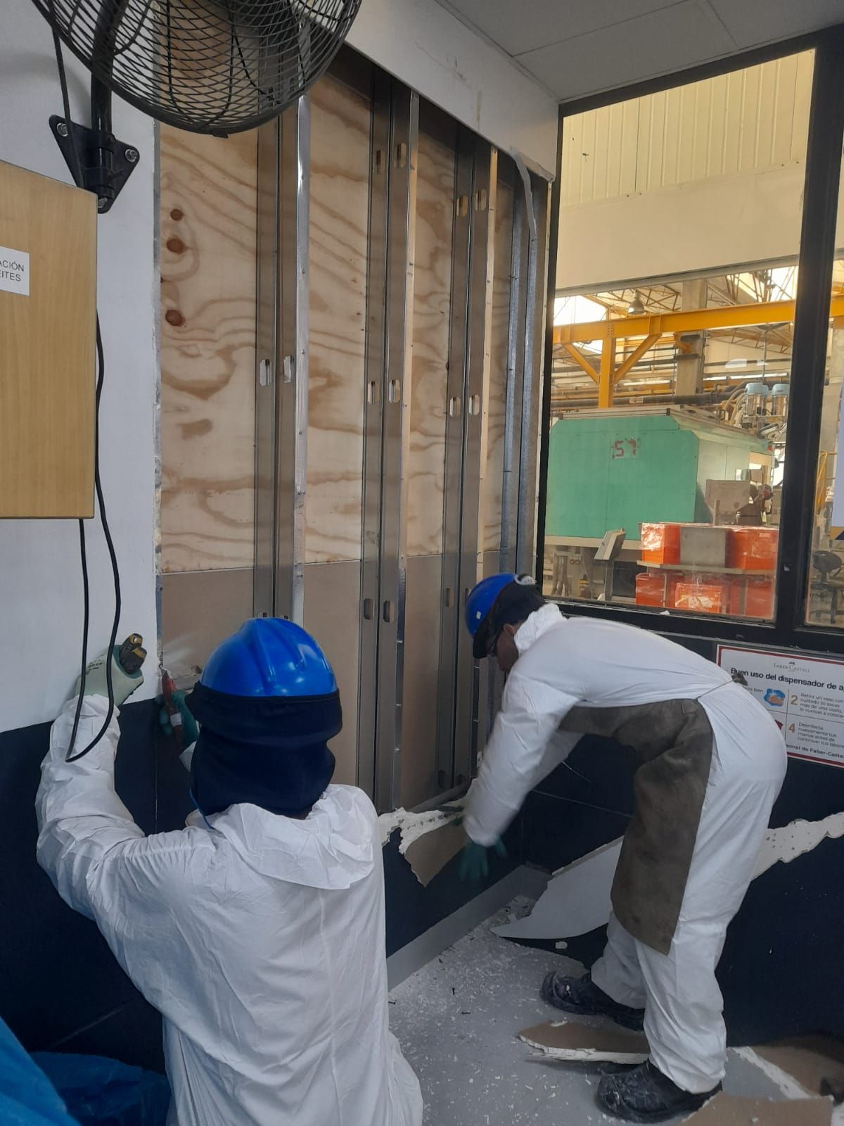 Dos trabajadores con trajes de protección instalan aislamiento de pared en una habitación con vistas a una fábrica.