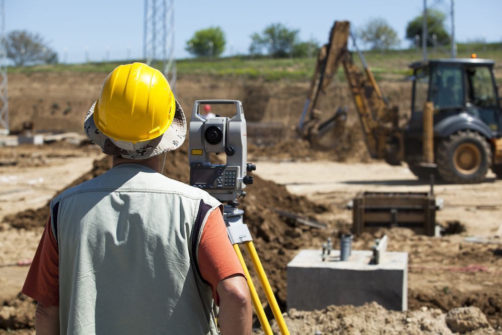 A worker wearing a yellow hard hat looks through a surveying instrument at an outdoor construction site with machinery — Jim Anderson Earthmoving Pty Ltd In Coopernook, NSW.