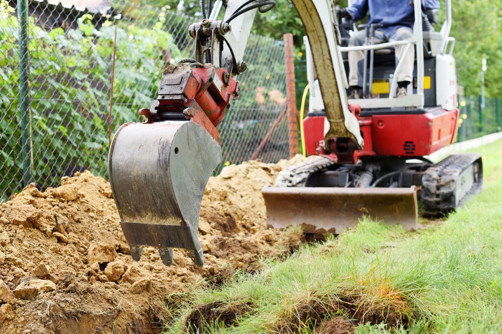 A compact excavator digs a narrow trench in a grassy yard near a chain-link fence — Jim Anderson Earthmoving Pty Ltd in Coopernook, NSW.