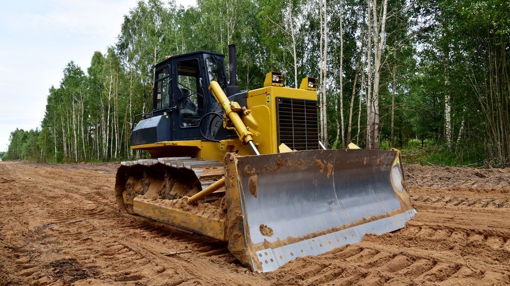 A yellow bulldozer sits on a dirt path in front of a forest, with fresh tracks visible in the soil  Jim Anderson Earthmoving Pty Ltd in Coopernook, NSW.