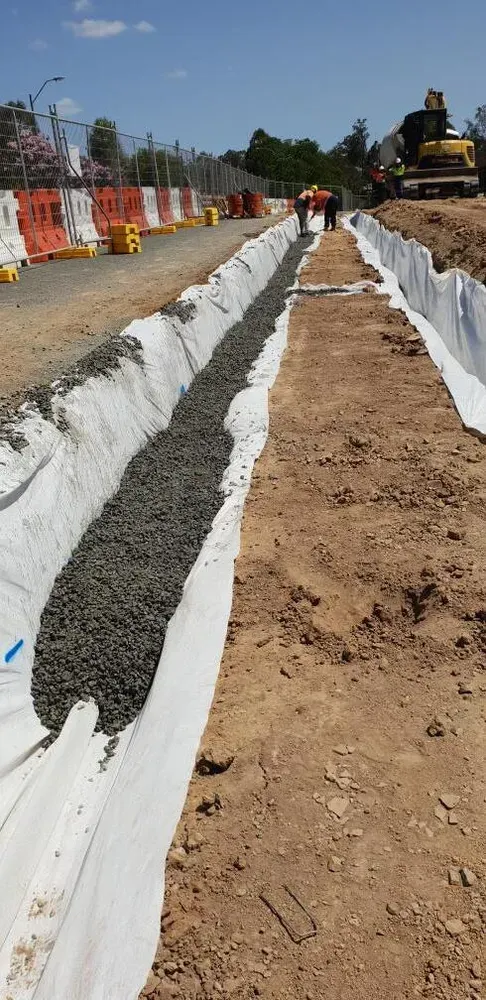 A narrow trench lined with white fabric and partially filled with gray gravel at a construction site.