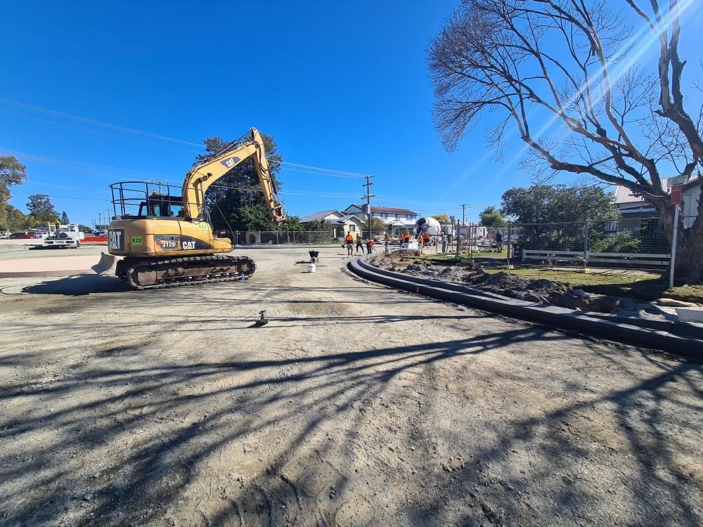 A yellow excavator stands on a dirt construction site near a new concrete curb under a clear blue sky — Jim Anderson Earthmoving Pty Ltd in Coopernook, NSW.