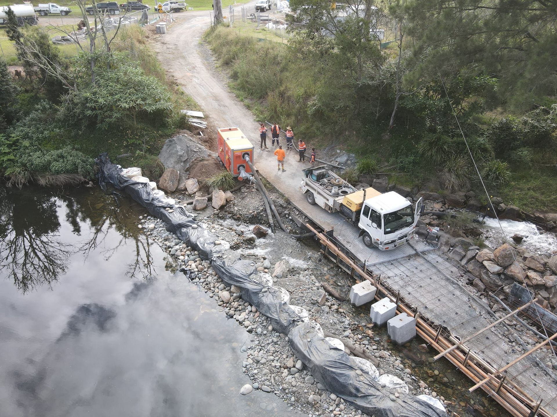 Construction site with a truck on a temporary bridge over a river; workers in orange vests nearby — Jim Anderson Earthmoving Pty Ltd In Coopernook, NSW