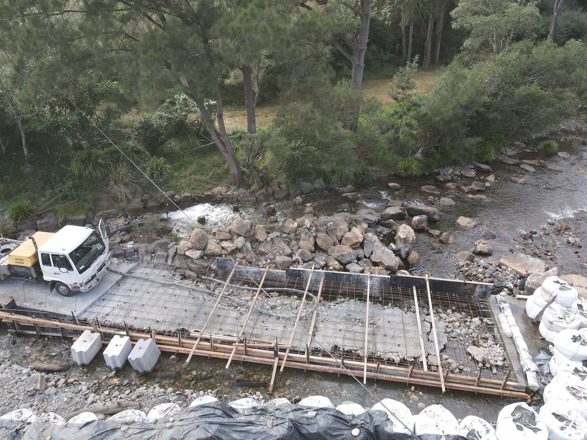 Construction site in a creek bed: concrete foundation with rebar, rocks, small truck, and surrounding greenery — Jim Anderson Earthmoving Pty Ltd In Coopernook, NSW