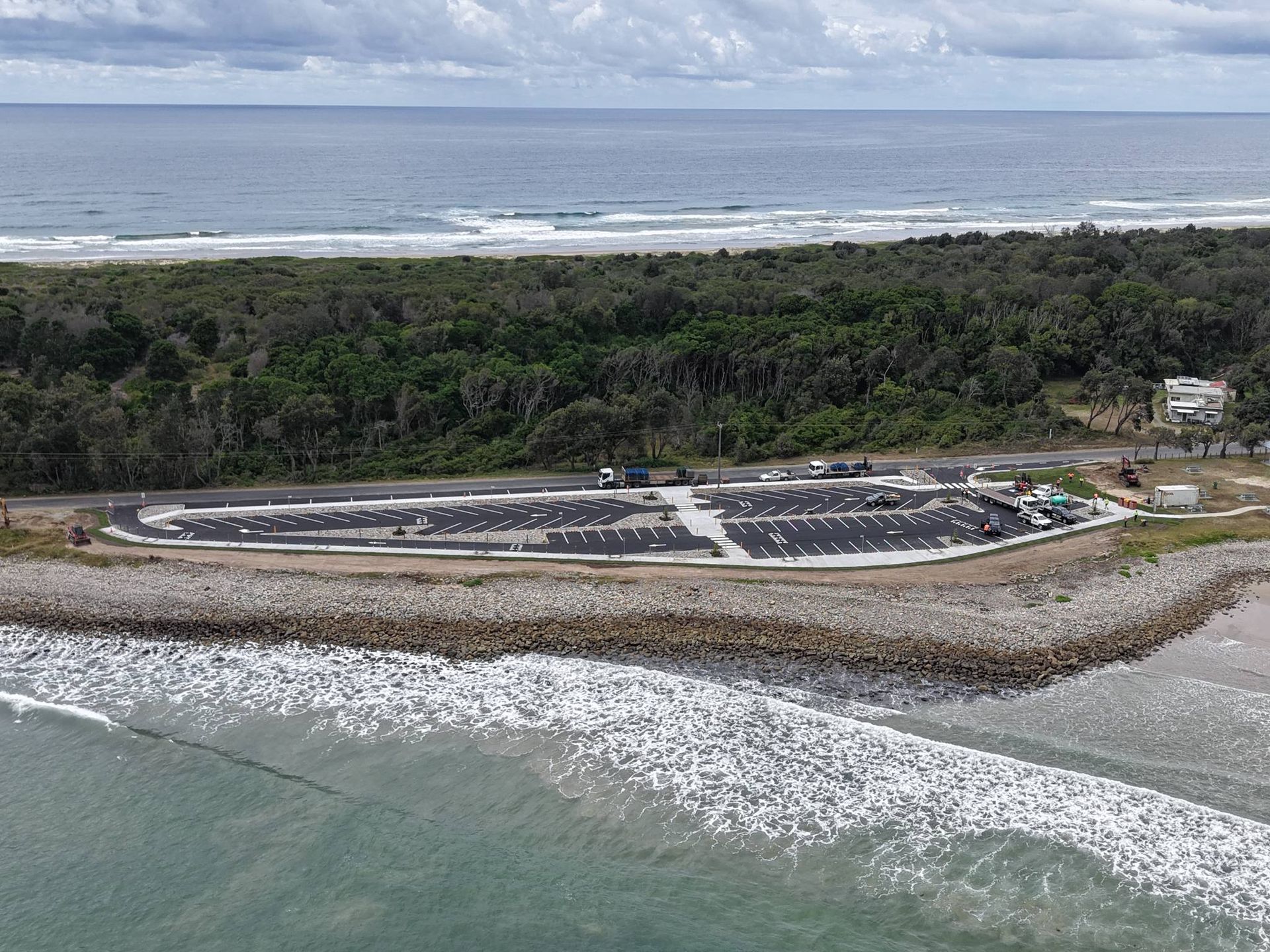 Aerial view of a coastal car park, rocky breakwater, beach, and dense green trees against the ocean and cloudy sky — Jim Anderson Earthmoving Pty Ltd In Coopernook, NSW