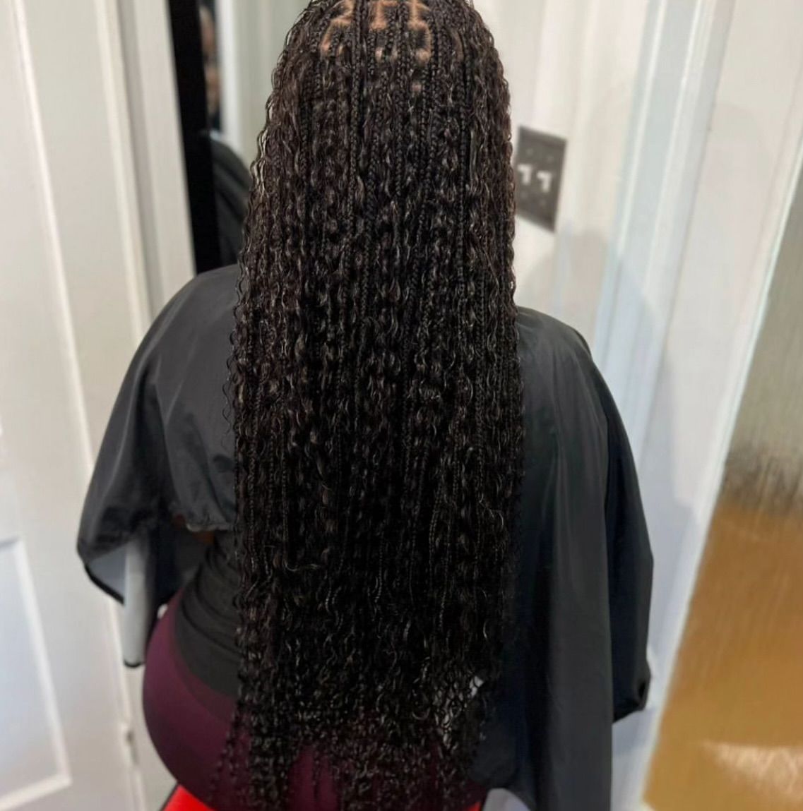 Woman with long, dark, wavy braids, wearing a black salon cape, sitting.