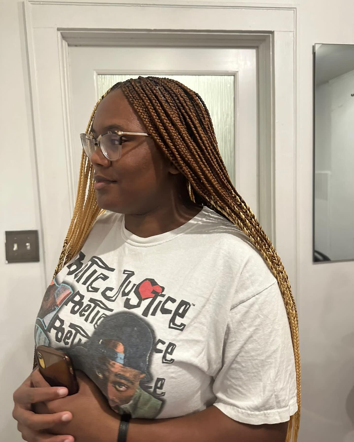 Woman with long, light brown braids, glasses, and a graphic tee. Holding a phone, looking slightly to the side.