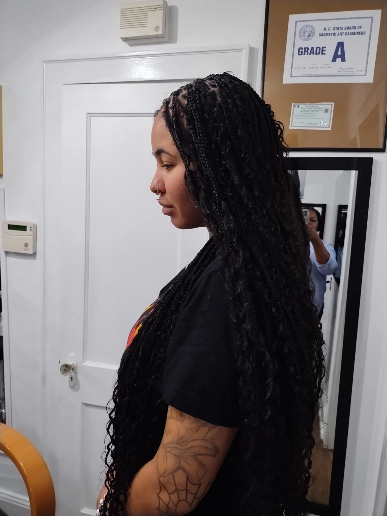 Woman with long, black braids stands in a room with a white door and mirror. She has tattoos on her arm.