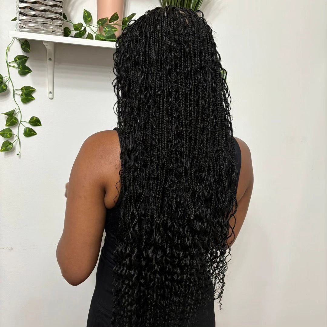 Woman with long, black micro braids; standing in front of a white wall with greenery.
