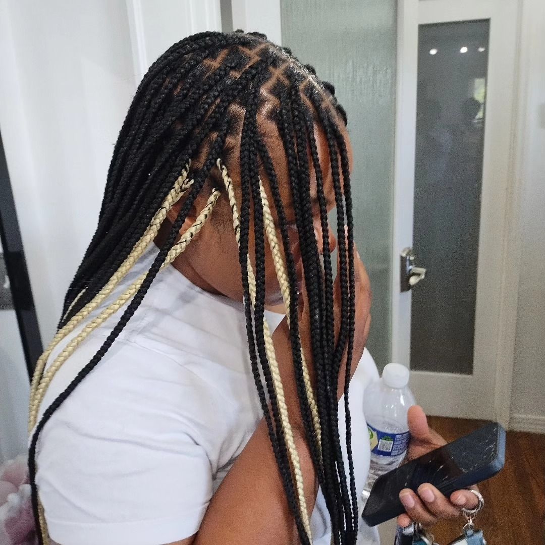 Woman with box braids, some blonde, holding phone. Indoors, wearing white shirt, looking down.