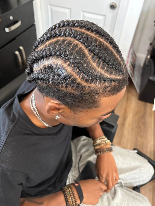 Man with wavy cornrow braids and faded sides, wearing jewelry, sitting.