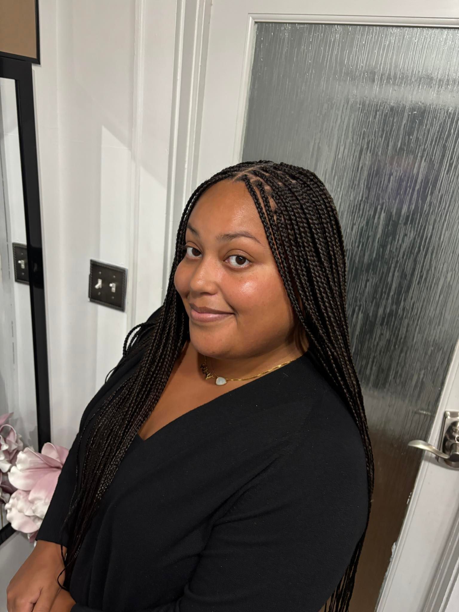 Woman with long box braids smiles, wearing black top, in front of a white door.