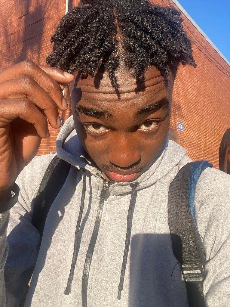 Young Black man with dreadlocks, wearing a gray hoodie and backpack, looking at the camera. Brick wall background.