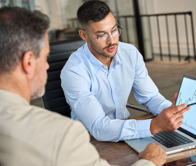 Two men are sitting at a table looking at a laptop computer.