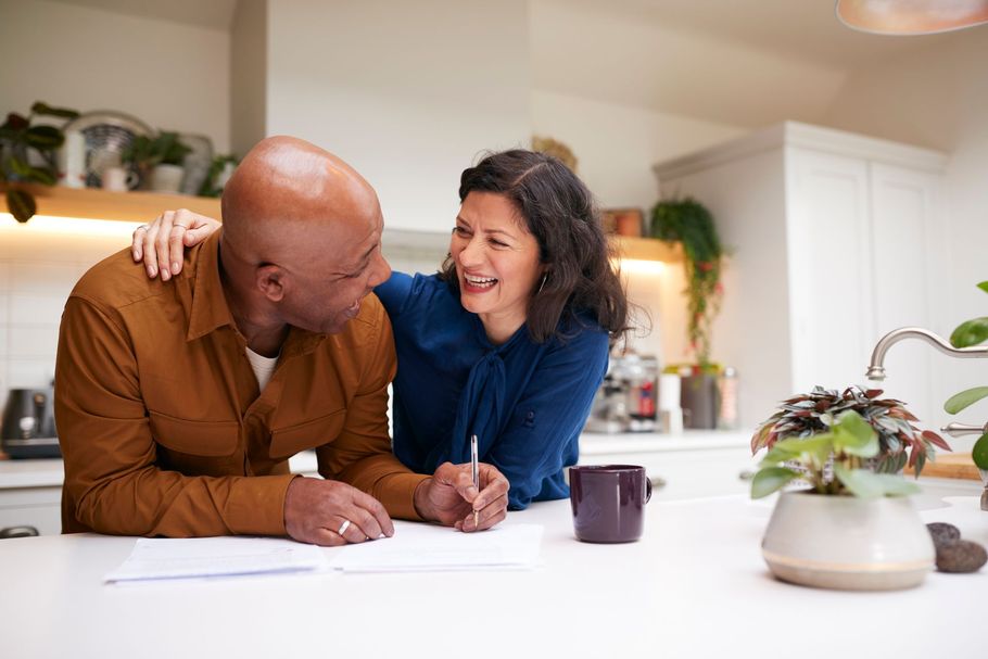 Two people smiling and leaning over a white kitchen counter with a mug and potted plant.