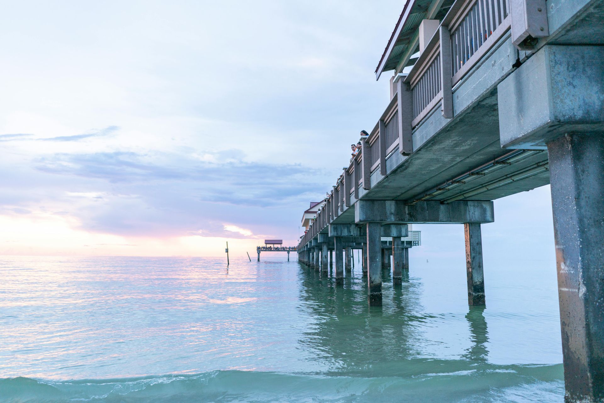 Seaside pier at sunset, with concrete supports over calm turquoise water and a pastel sky