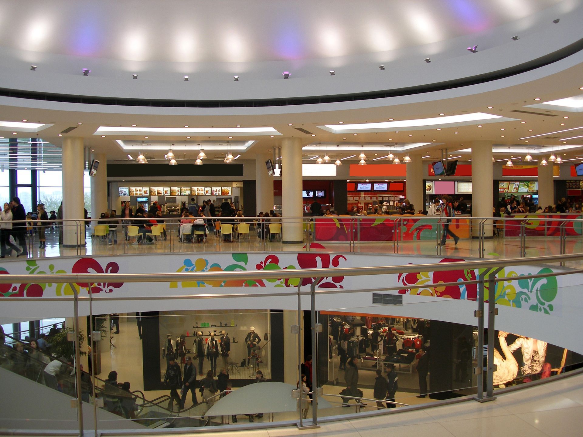 Multi-level mall interior with shops, food court, and people. White walls, colorful accents, overhead lighting.